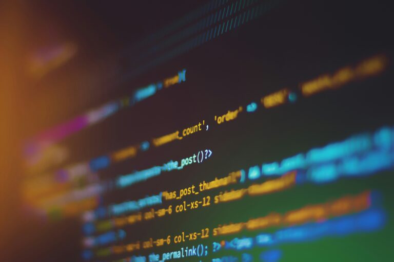 A close-up image of colorful lines of code displayed on a computer screen, representing programming or software development. The code is slightly blurred, with hues of blue, orange, and green, creating an abstract and vibrant visual effect.